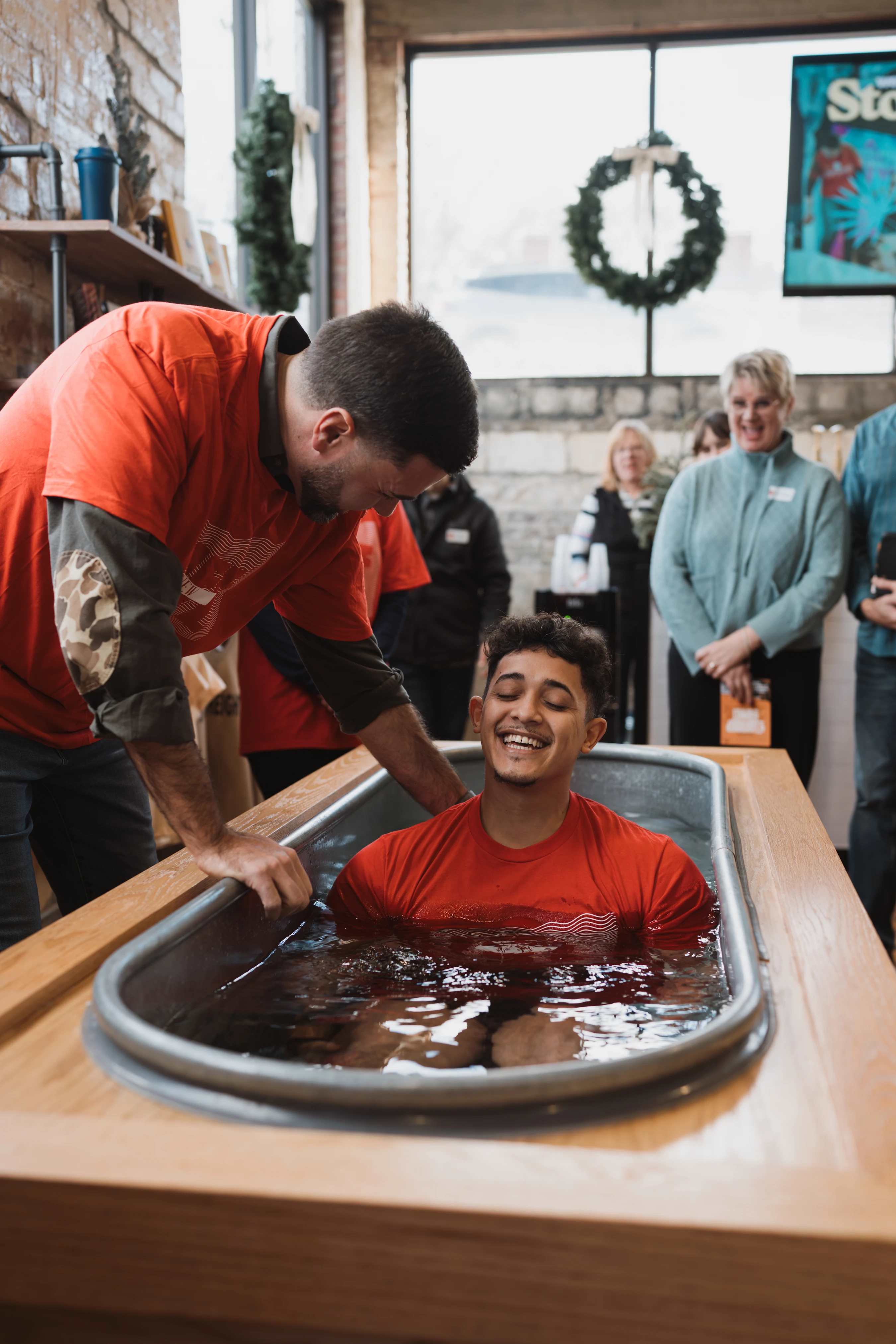 A young man in a red t-shirt smiles with his eyes closed while sitting in a metal tub of water as another man baptizes him in front of smiling onlookers.